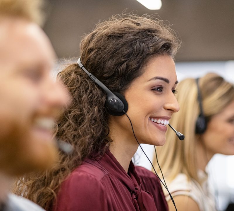 Smiling woman call center operator doing her job with a headset while working on computer. Positive smiling agents in conversation with customer over headset, sitting in row. Consulting and assistance helpdesk service.